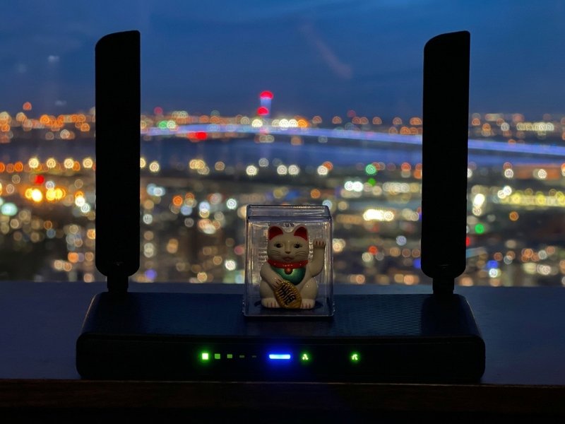 The MikroTik router this site runs on, photographed at night with a waving lucky-cat charm on top and city lights glowing out of focus behind it.